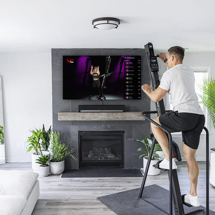 Male using the VersaClimber Home Climber with a live square display, engaging in a high-intensity vertical cardio workout with real-time metrics and guided on-demand fitness classes shown on the built-in tablet screen in his livingroom