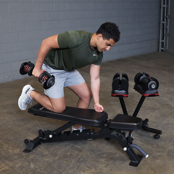 A person performing a single-arm tricep kickback on a weight bench with a Body-Solid Tools SDBX132 Adjustable Dumbbell, while another adjustable dumbbell rests on a storage stand nearby.
